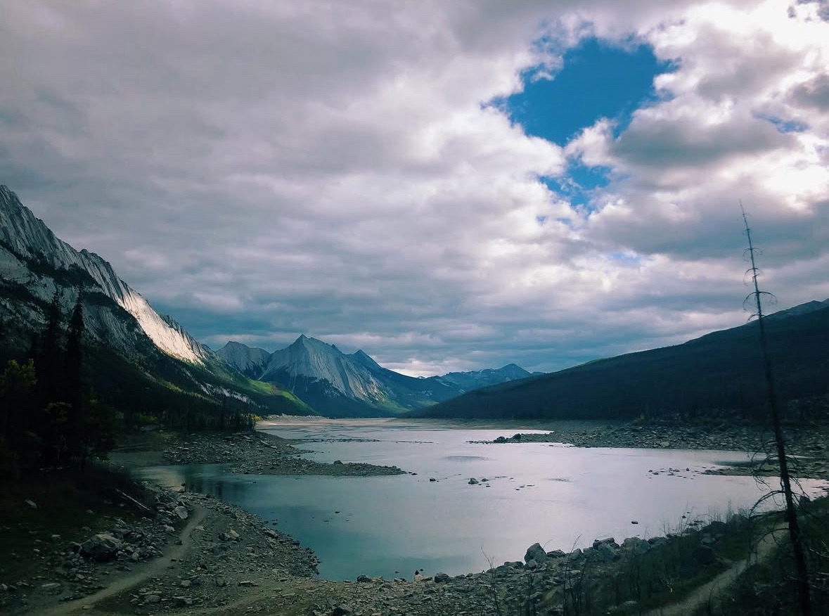 Under a mostly cloudy sky with a few patches of blue is a still blue lake sitting in a rocky valley bed between mountains, some sloping and others with more dramatic, sharp cliffs, spotted with sunlight.