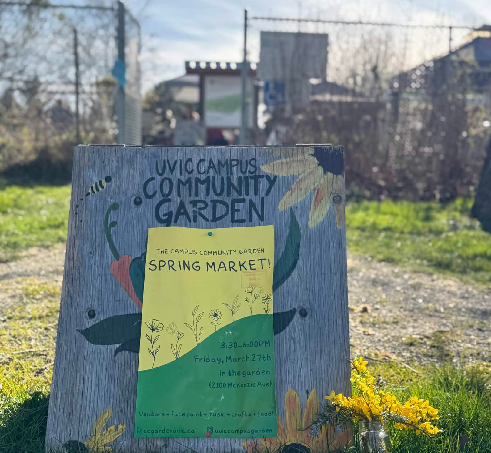 A wooden sandwich board with painted flowers and vegetables on it reads "UVic Campus Community Garden" and has a poster pinned to it, yellow and green, advertising the Spring Market.